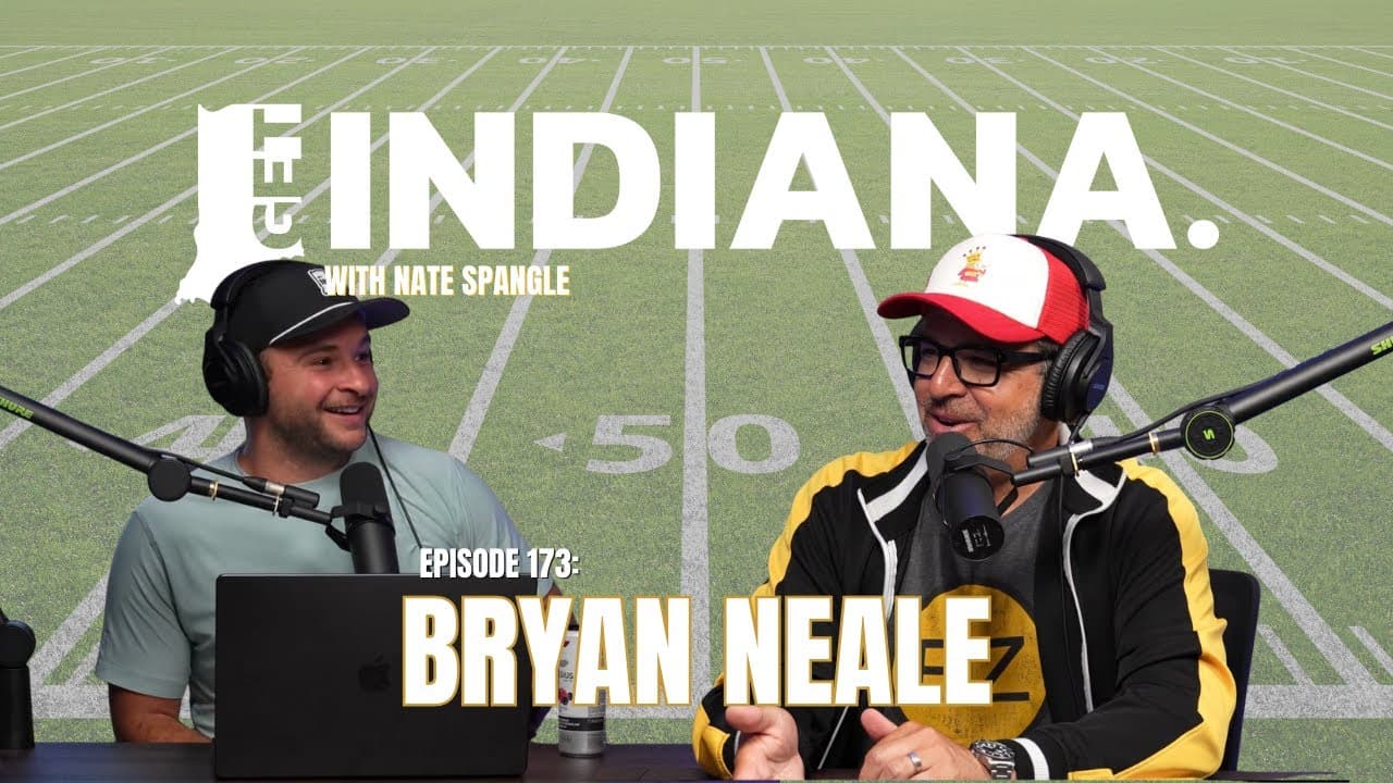 Two men in headphones discuss business on a podcast set, with a football field backdrop in Indiana.
