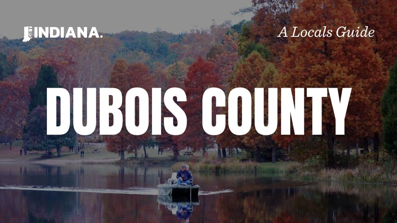 A serene lake in Dubois County, Indiana, surrounded by vibrant autumn trees and a lone fisherman in a small boat.